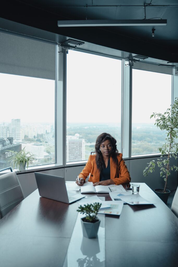 Mulher negra, com blaser laranja, sentada em uma sala de reunião corporativa, com vista para a cidade.